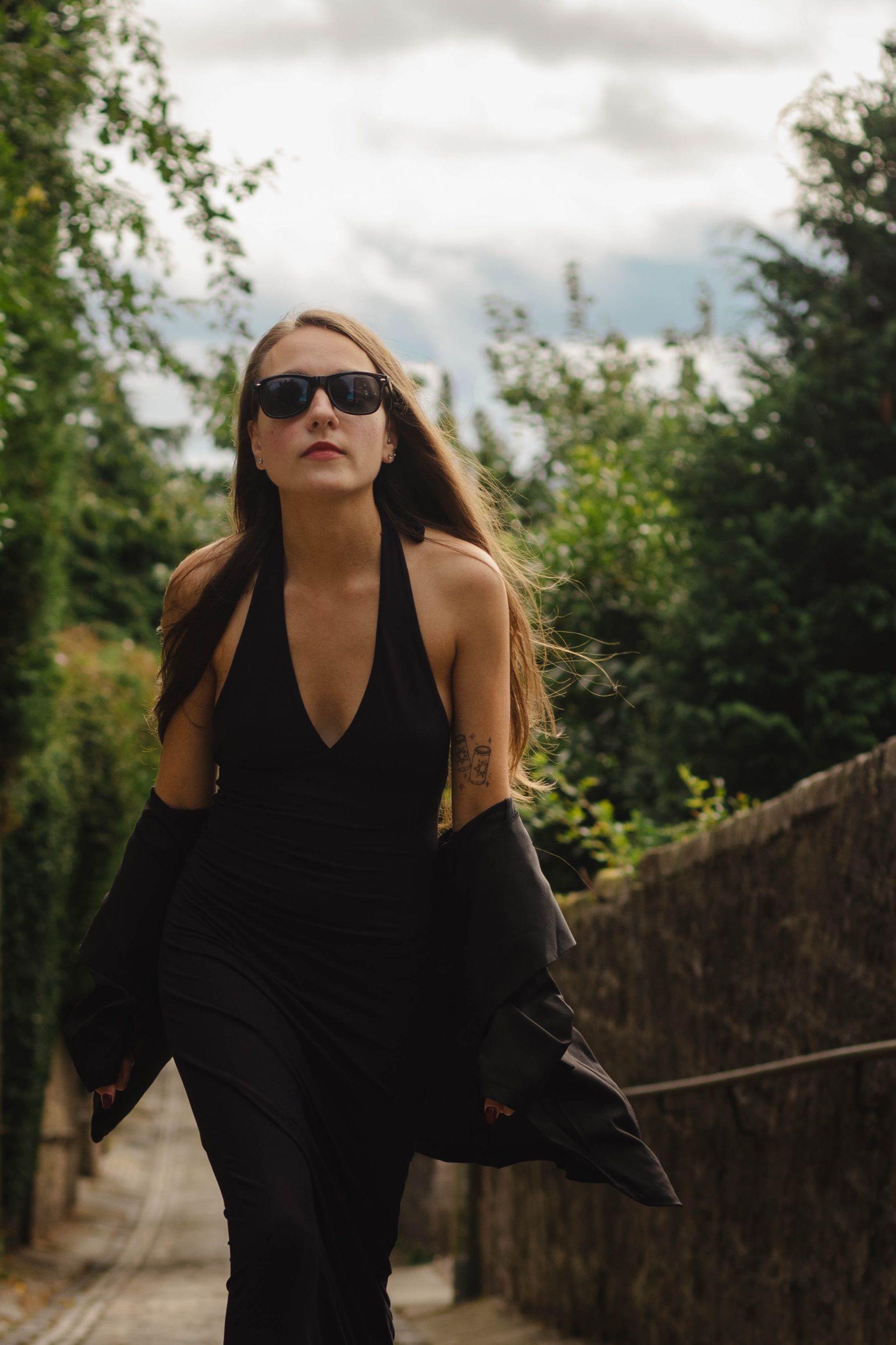 Woman in a black halter dress and sunglasses walking up a leafy lane