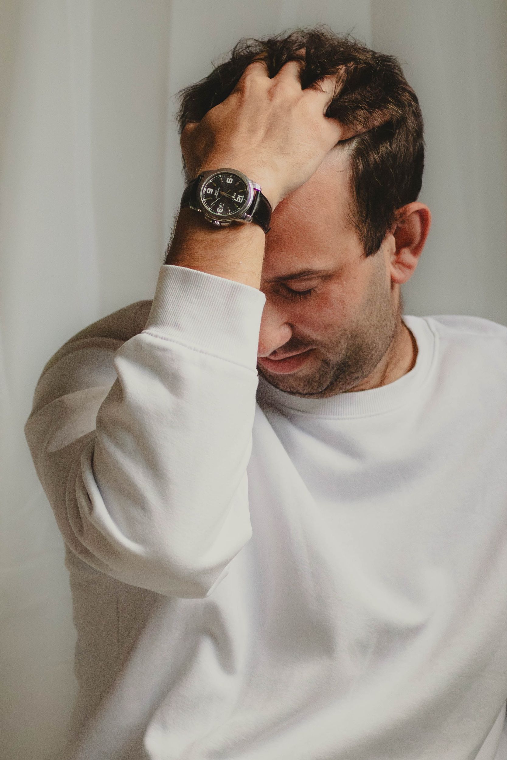 Man in a white sweatshirt running a hand through his hair against a pale curtain