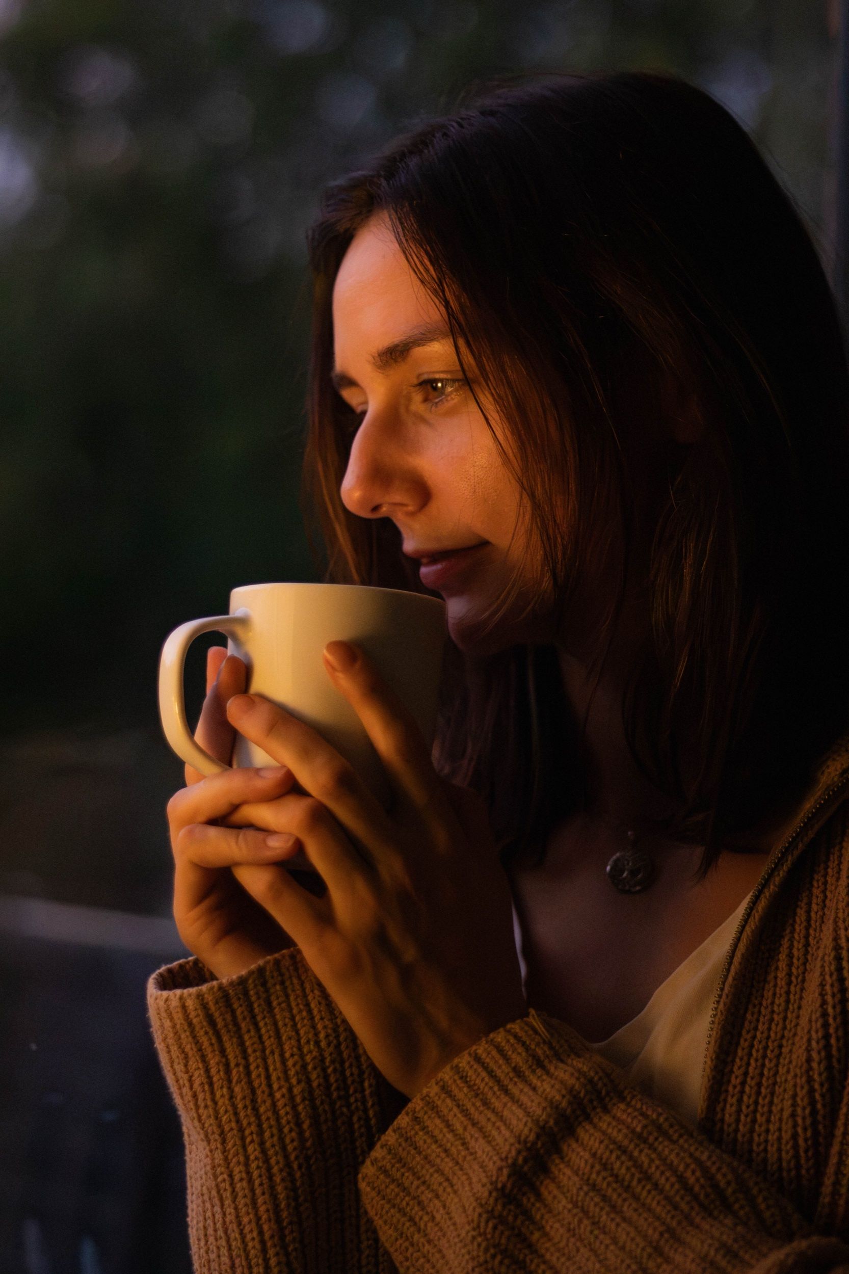 Woman in a knitted cardigan holding a mug to her lips in warm evening light